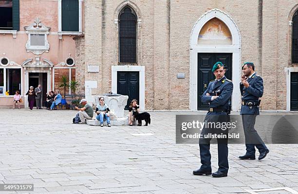 Ein Platz in Venedig. Polizisten gehen Streife. Müde Touristen ruhen sich am Brunnen aus.