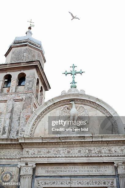 Eine Möwe fliegt über eine weiße Kirche. Auf der Fassade steht "DIVO IOANNI APOSTOOI ET EVANGELISTAE PROTECTORI ET SANCTISSIAE CRVCI" gehauen.