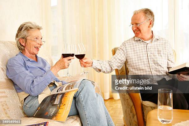 Bonn: Living in old age: Seniors couple in living room at home. Reading books, playing Sudoku and drinking red wine.