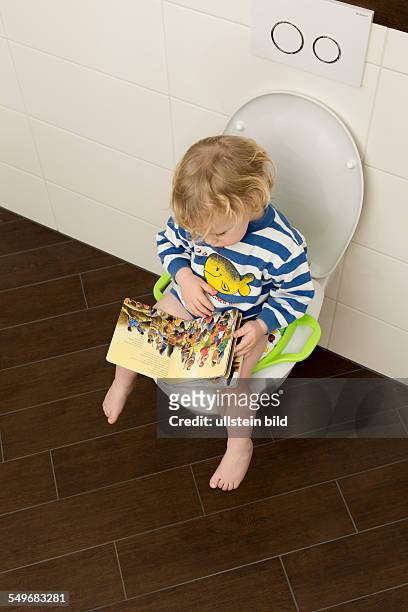 Year old boy with childrens seat and childrens book on a toilet.