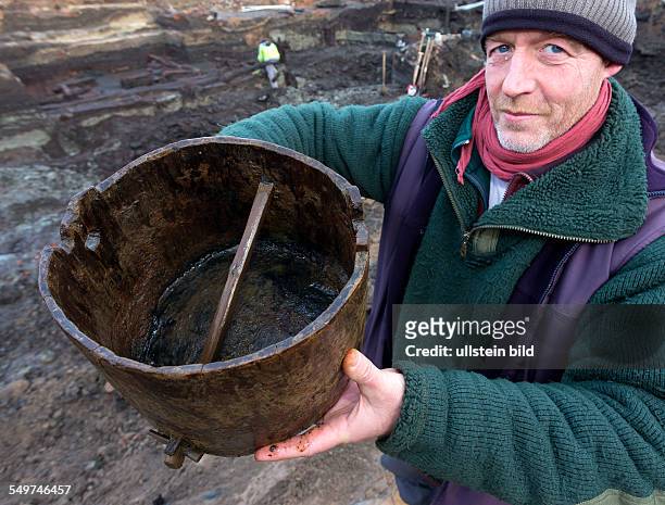 Grabungsleiter zeigt das gefundene Schoepfgefaess aus Eiche vor der Ausgrabung der grossflaechigen Holzlage aus der Zeit vor 1250 in der Greifswalder...