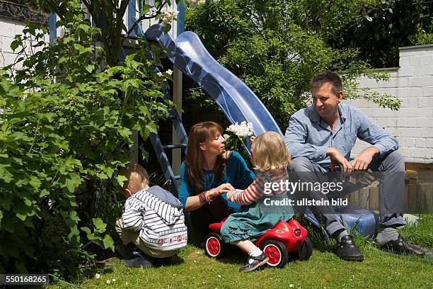 Scene family: father, mother and two children in the garden. It's a warm spring day. The father is sitting on a slide, the little girl playing with...