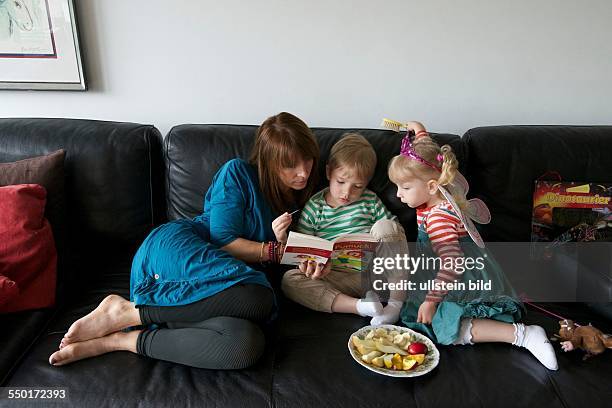 Family scene: A mother sits with her young son and her little daughter on the couch. They read together a children's book. To snack is a plate of...