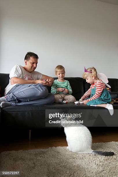 Family Scene: The father, a little boy and a little girl sitting on the sofa in the living room and play with surprise eggs. Front of the sofa sits a...