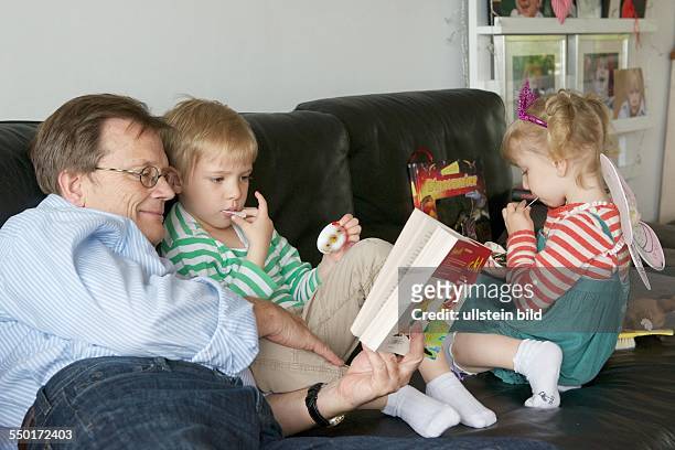 Family Scene: The Grandfather reads a little boy and a little girl from a children's book in front of a story. Grandfather and grandchildren are...