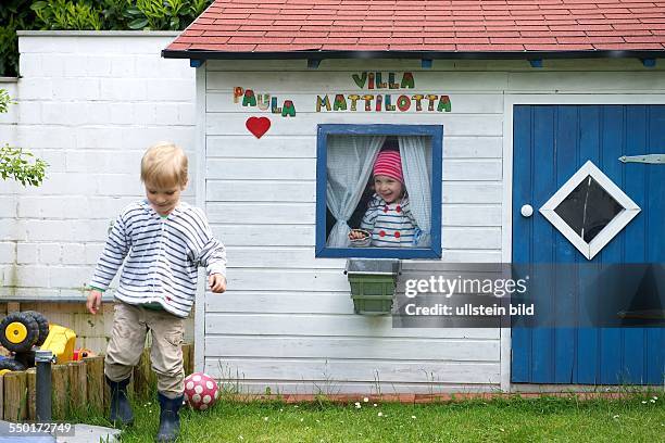 Family Scene: A little boy and a little girl playing in the garden. The girl looks out the window of a brightly painted summer house. On the wall is...