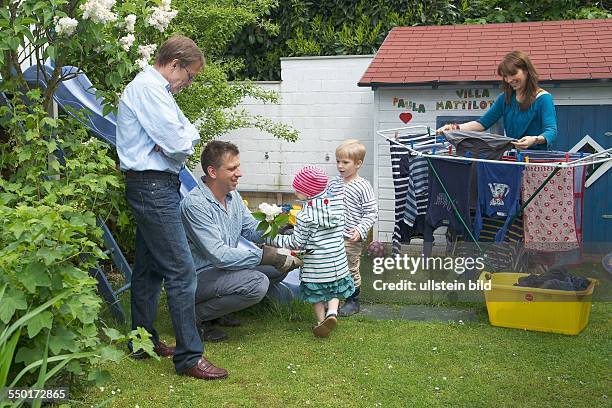 Family Scene: The father plays with the two children in the garden. The mother hangs freshly washed clothes to dry on a clothes line. The grandfather...