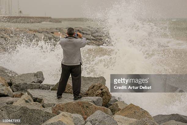 Man taking pictures with smartphone during heavy storm with waves crashing on the rocks. Maresme, Catalonia, Europe.
