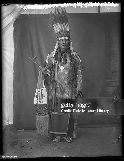 Portrait of an unidentified Native American Arapaho man with feather headdress, Ghost Dance shirt or dress, and holding a pipe and beaded bag at the...
