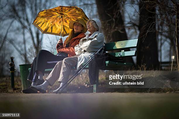 Zwei ältere Damen , geniessen die Vorffühlingshaften Temperaturen unter dem Schutz eines Schirmes auf einer Parkbank im Botanischen Garten in Berlin