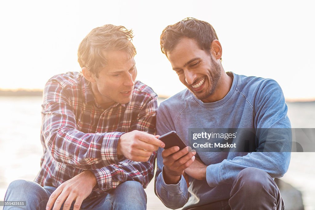 Happy male friends reading text message on mobile phone against sea