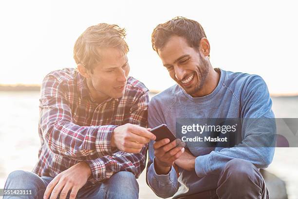 happy male friends reading text message on mobile phone against sea - male cellphone stock-fotos und bilder