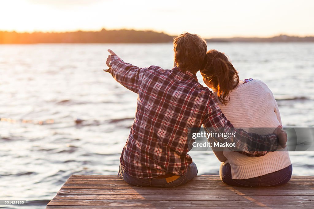 Rear view of man showing something to woman while sitting on pier against sea at sunset