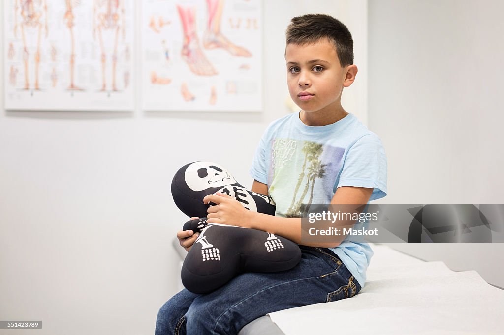 Portrait of boy holding skeleton stuffed toy on examination table at orthopedic clinic