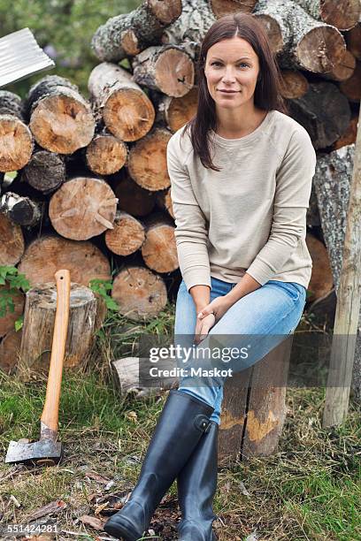 full length portrait of woman sitting on tree stump against stacked firewood at yard - benen over elkaar geslagen stockfoto's en -beelden