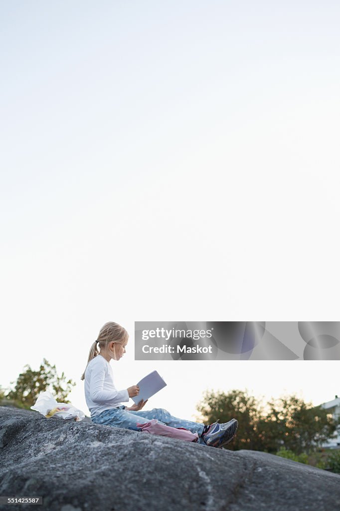 Full length side view of girl doing homework while sitting on rock against clear sky