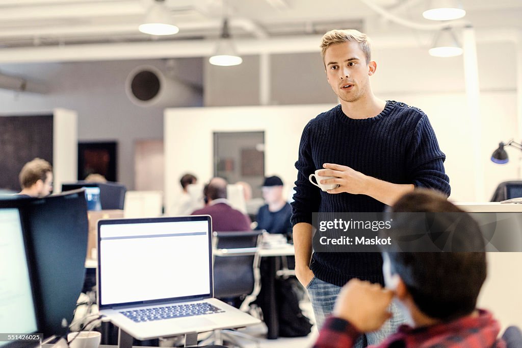 Businessman holding coffee cup while discussing with colleague in office