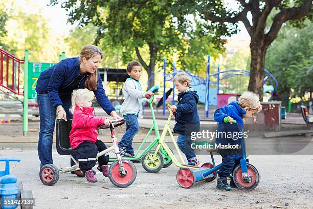 female teacher assisting girl to ride tricycle on playground - kinderopvang stockfoto's en -beelden
