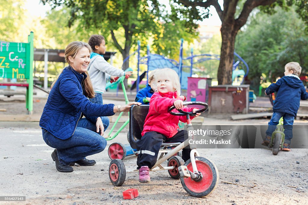 Happy female teacher assisting girl to ride tricycle on playground