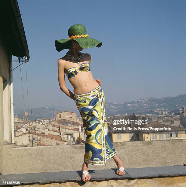 Sixties Fashion - A young female model wears a matching bikini and zulu style skirt in printed cotton of blue, yellow and green with a large green...