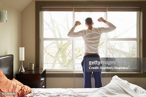 rear view of young man stretching in front of bedroom window - morning bed stretch stock pictures, royalty-free photos & images
