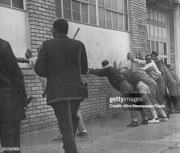 During a riot, police officers with billy clubs guard suspected looters, who stand with their hands against a brick wall at the corner of Howard and...