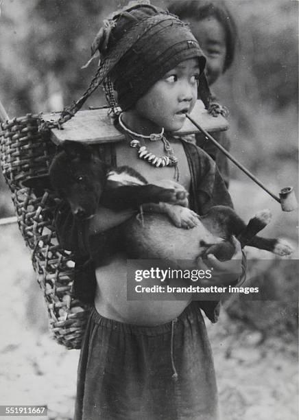 Border area between Thailand and Burma . 10-year-old girl from the Akha with pipe and dog and provided with a shoulder board and headband carrying...