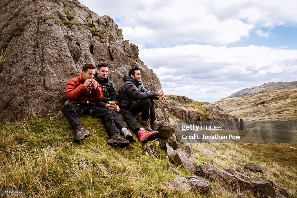 Three young male hiking friends eating sandwiches, The Lake District, Cumbria, UK