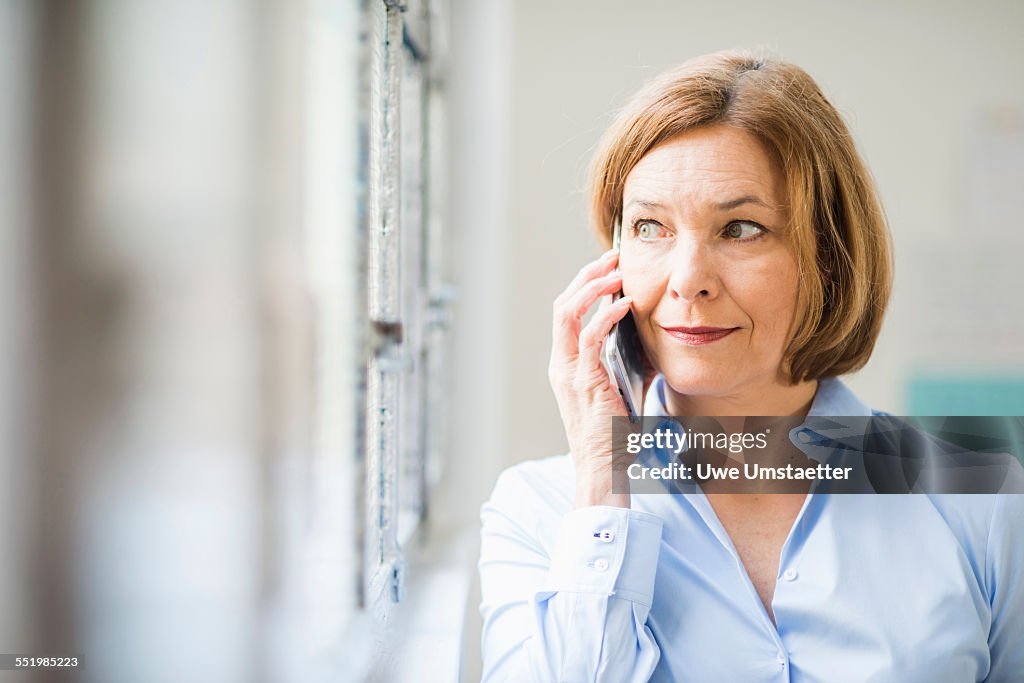 Mature businesswoman looking out of office window and chatting on smartphone