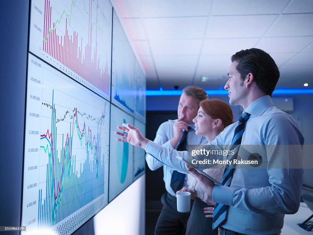 Three business people discuss graphs on screen in meeting room
