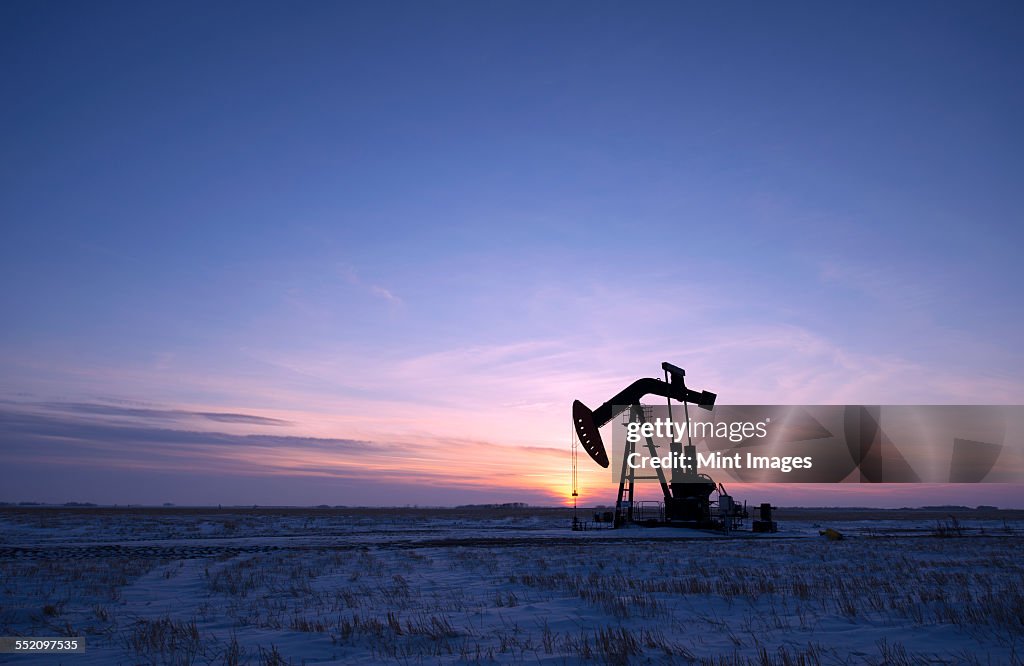 An oil drilling rig and pumpjack on a flat plain in the Canadian oil fields at sunset.