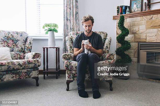 young man sitting at home with small white kitten - armstoel stockfoto's en -beelden