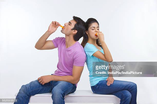 young couple eating ice lollies while sitting on bench over white background - wassereis mit orangengeschmack stock-fotos und bilder