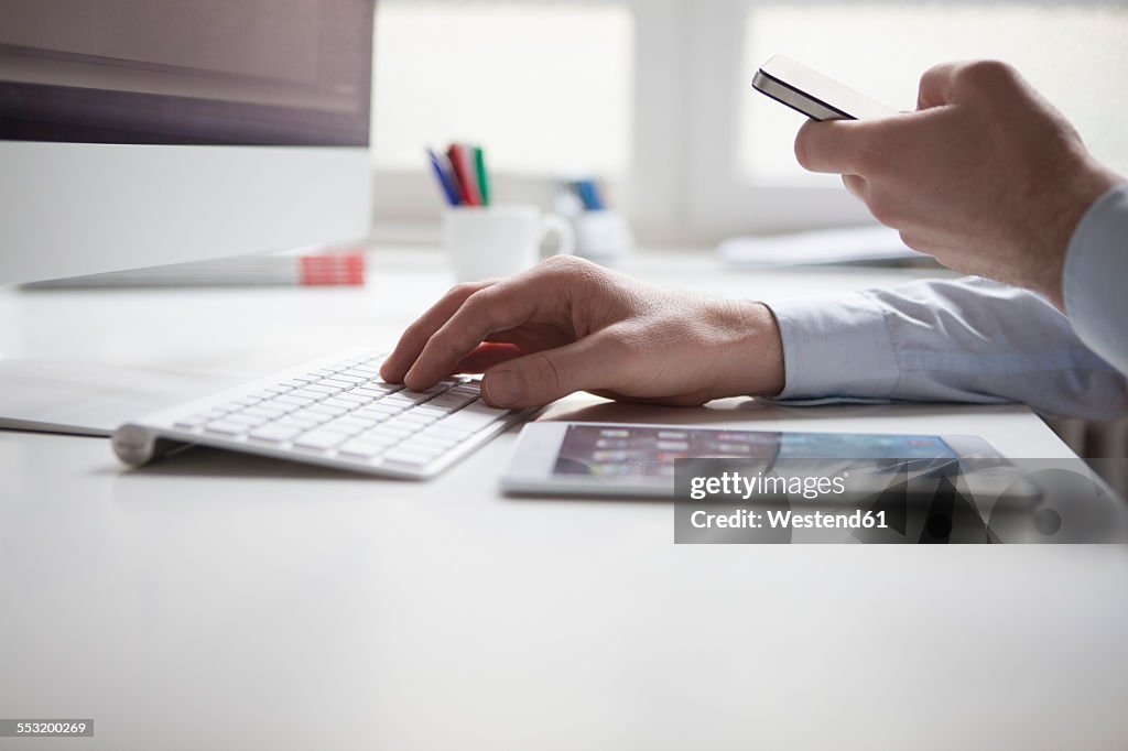 Man's hand typing on keyboard of computer while using smartphone