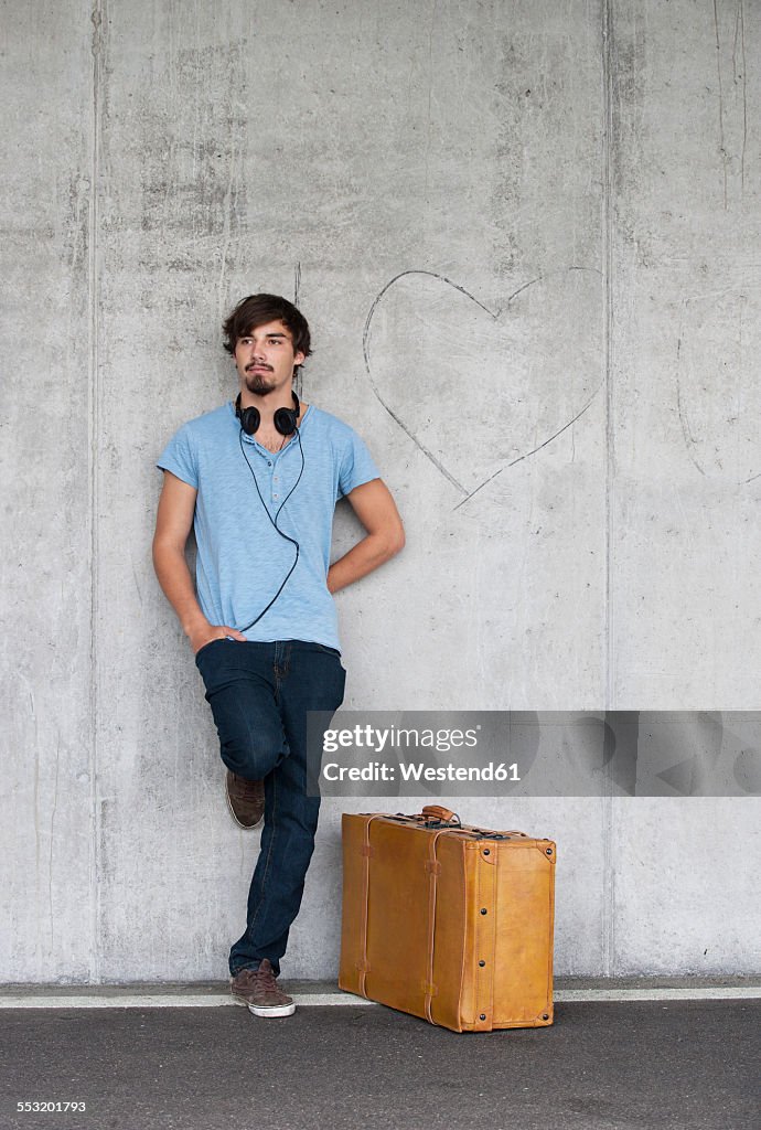 Man with leather suitcase and headphones leaning at concrete wall