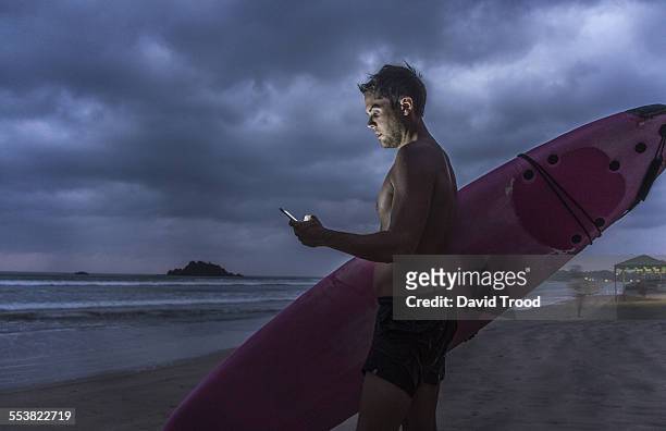 man with surfboard using smart phone at dusk - beach hold surfboard stock-fotos und bilder