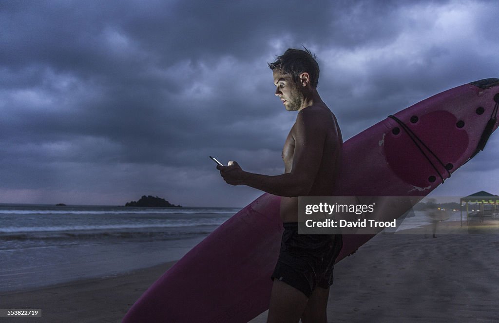 Man with surfboard using smart phone at dusk