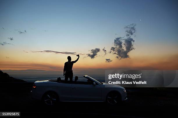 man taking photo of sunset, standing in open car - on the move rear view stockfoto's en -beelden