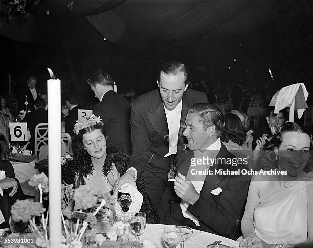 Actor Errol Flynn sits at a table as actor Basil Rathbone pours champagne for him with Veronica Cooper and actress Dolores Del Rio during an event in...