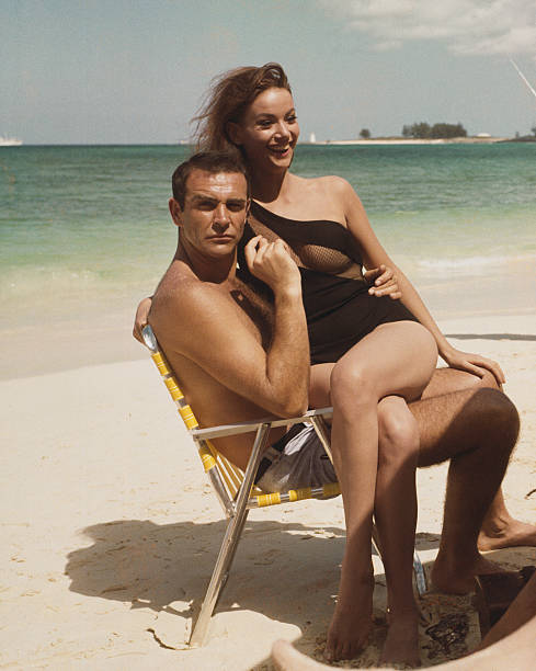 Scottish actor Sean Connery and French actress Claudine Auger pictured together wearing bathing costumes and sitting on a folding chair on a beach in...
