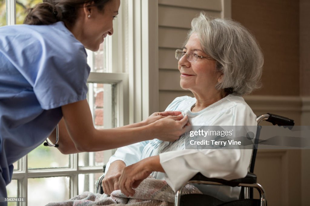 Nurse buttoning shirt of patient in wheelchair near window