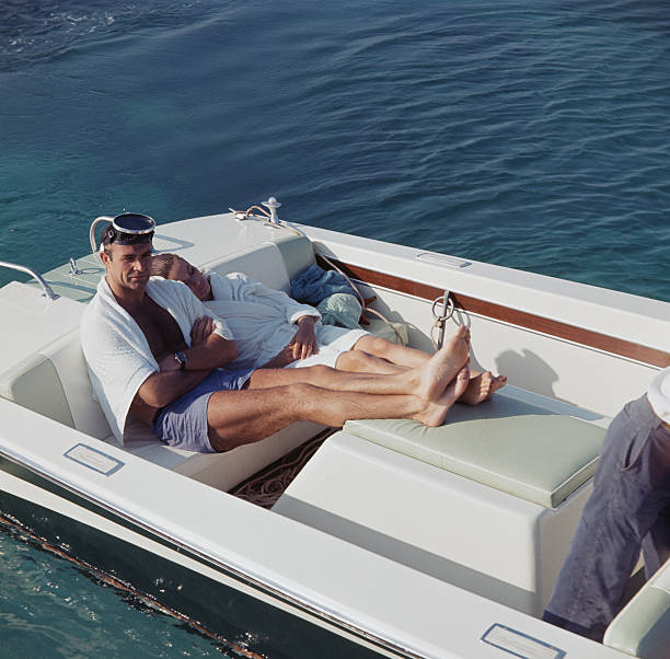 Scottish actor Sean Connery pictured sitting in the back of motor launch wearing blue shorts, white towel and scuba mask with French actress Claudine...