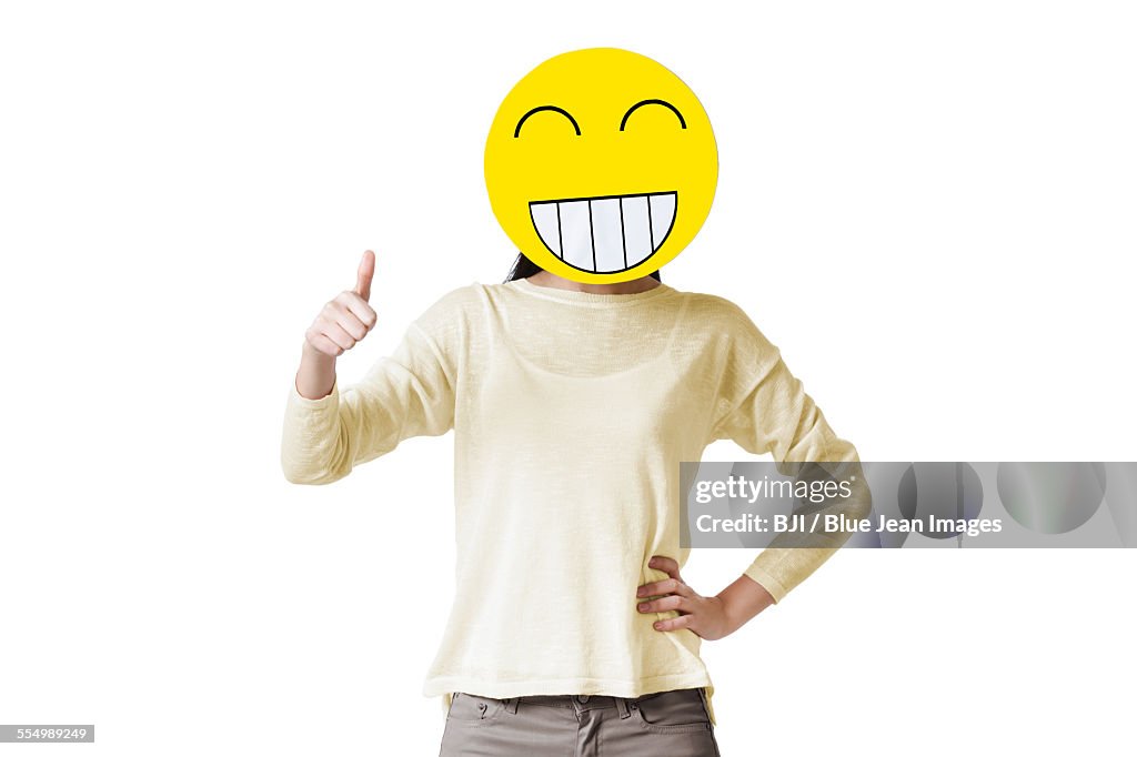 Young woman giving thumbs up with happy emoticon face in front of her face