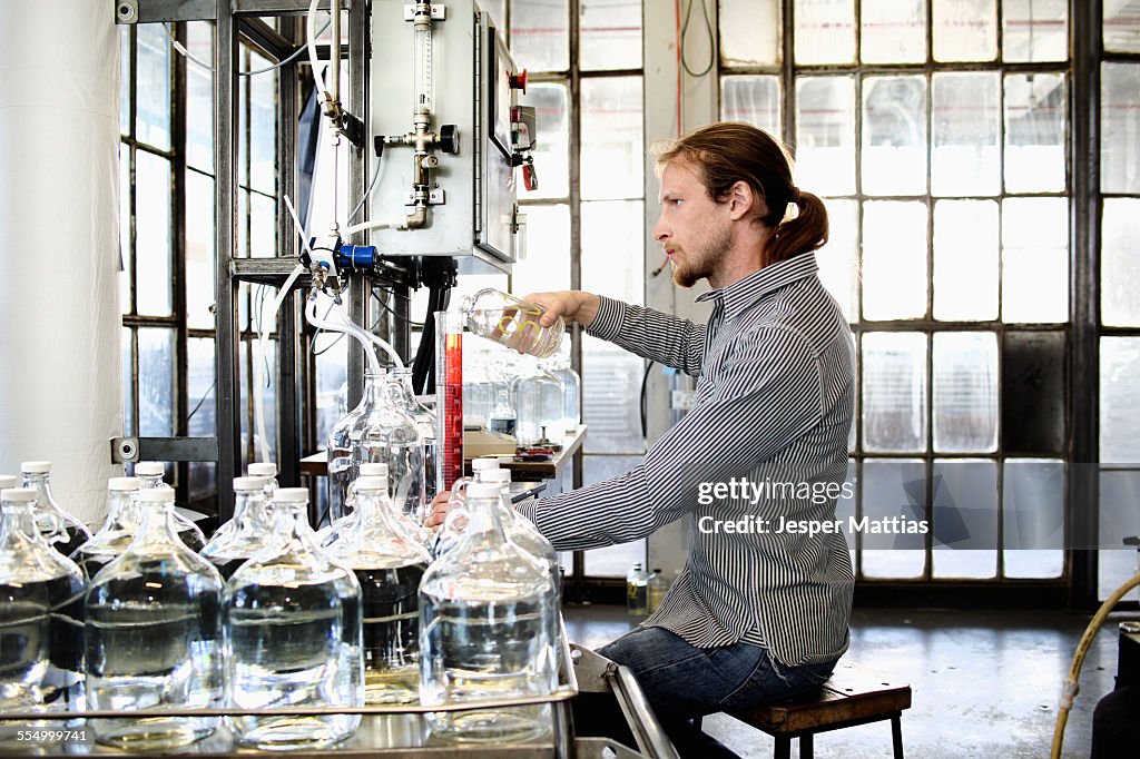 Young male vodka distiller pouring liquid into cylinder in distillery workshop