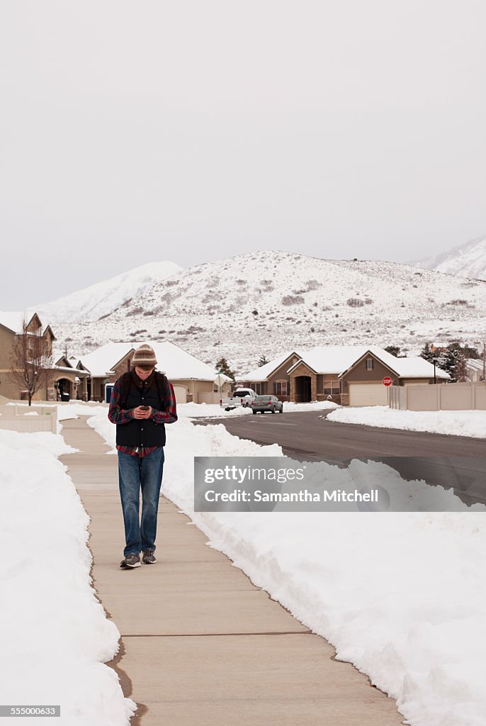 Teenage boy reading smartphone texts whilst walking path in snow covered village