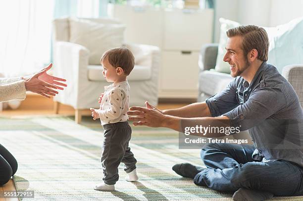 happy parents helping little son (2-3 years) walking in living room - first steps stock-fotos und bilder