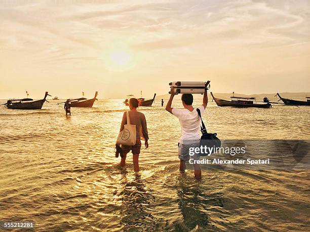 Travelling couple in Thailand walking in the ocean to the longtail boat with their luggage bags above the head