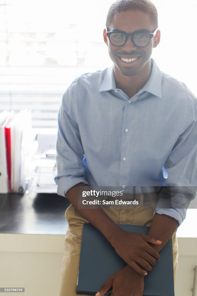 Portrait of smiling young man wearing glasses and blue shirt leaning on desk in office