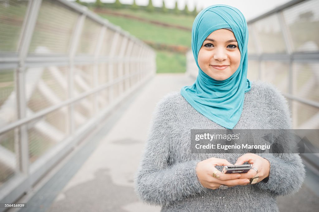Portrait of young woman wearing turquoise hijab using smartphone on footbridge