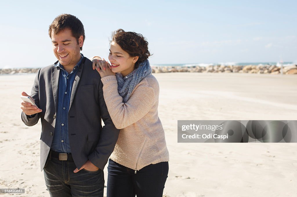 Young couple reading smartphone texts on beach, Tel Aviv, Israel